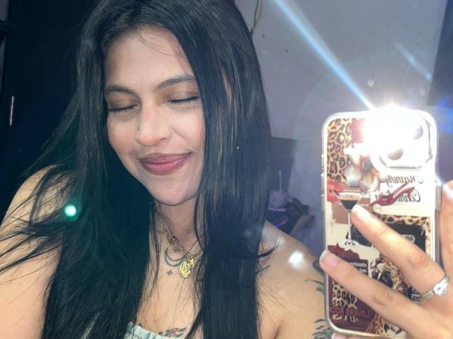 NyraVanel, eine lächelnde Frau mit langen, schwarzen Haaren, macht ein Selfie mit einem Handy, das mit Leopardenmuster und Anime-Designs verziert ist. Ihre Lippen sind leicht geöffnet, die Augen geschlossen, sie strahlt eine sexuelle Spannung aus..