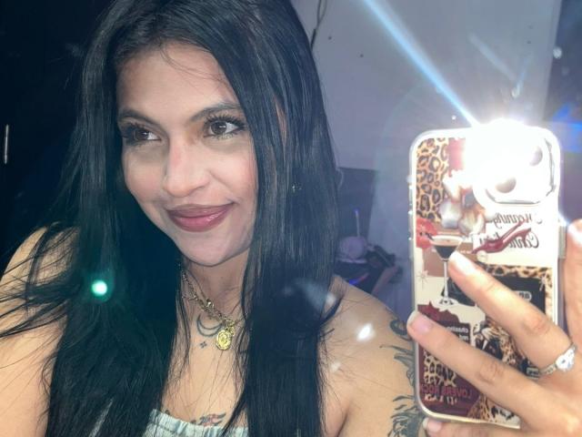 Close-up selfie of NyraVanel with long black hair, smirking, holding a phone with a leopard print case, illuminated by a flash. She has a tattoo on her upper arm.