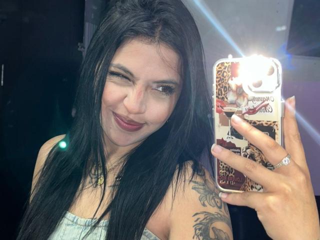 NyraVanel, a hot woman with long black hair and tattoos, takes a mirror selfie with a glowing phone. She's smirking, showing off her red lipstick and cleavage. Fucking sexy.