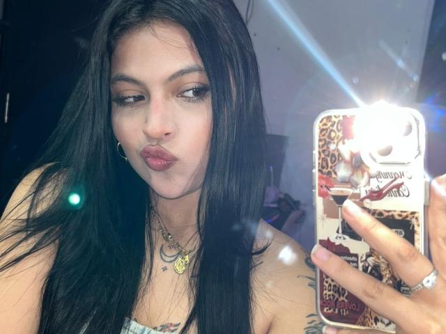 NyraVanel takes a selfie, holding a phone with a leopard print case, flashing a pouty expression. Her long black hair, slight makeup, and necklace are prominent. Bright flash in foreground.