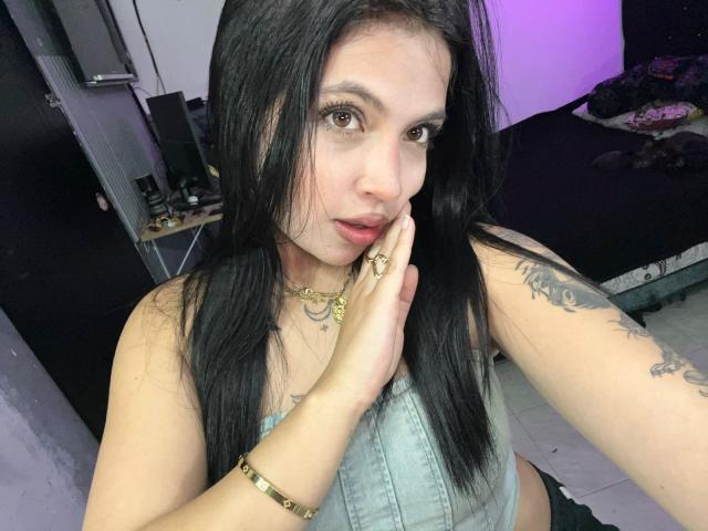 Close-up photo of NyraVanel, a woman with long black hair, pale skin, and tattoos on her arm. She's wearing a light blue tank top, touching her face with one hand. Her red lipstick stands out. Purple light in the background. Her expression is slightly seductive.