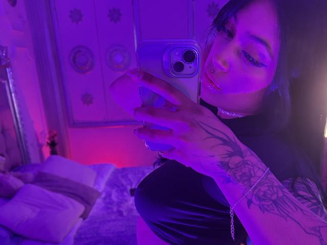 KatianPierce, con el brazo tatuado levantado, sosteniendo un teléfono, tomando un selfie en una habitación iluminada con luz púrpura, vistiendo una blusa negra, luz azul. Reflejándose en su rostro, una cama con almohadas al fondo..