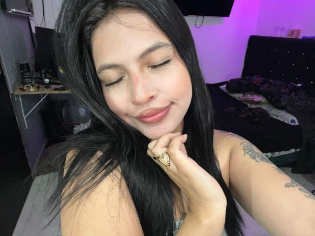 Close-up of NyraVanel, a Latina woman with long black hair, eyes closed, smiling, holding her chin with a gold ring, visible tattoos on her right arm, wearing a strapless top. Fucking cute.