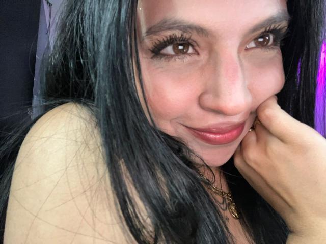Close-up foto van NyraVanel, een Latina vrouw met lang zwart haar, bruine ogen en rode lippenstift, die haar wang op haar hand, met een lichte glimlach, die een sexy en zelfverzekerde uitdrukking toont..