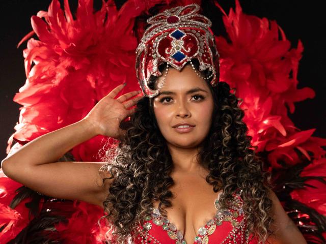 JedaZore, with curly black hair, wears a glittery red and silver headdress, and a low-cut red dress. Background filled with bright red feathers. Hand raised to head, sexy expression.