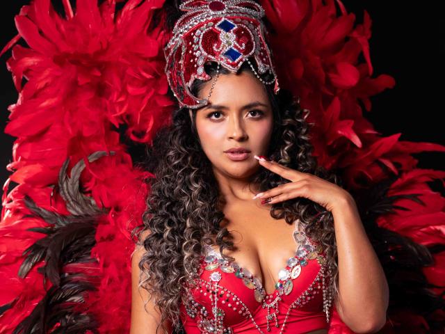 JedaZore, avec ses cheveux noirs et bouclés, porte une robe rouge ornée de sequins et un diadème élaboré. Des plumes rouges l'entourent, créant une tension sexuelle intense. Sa main effleure ses lèvres, dégageant un charme séduisant..