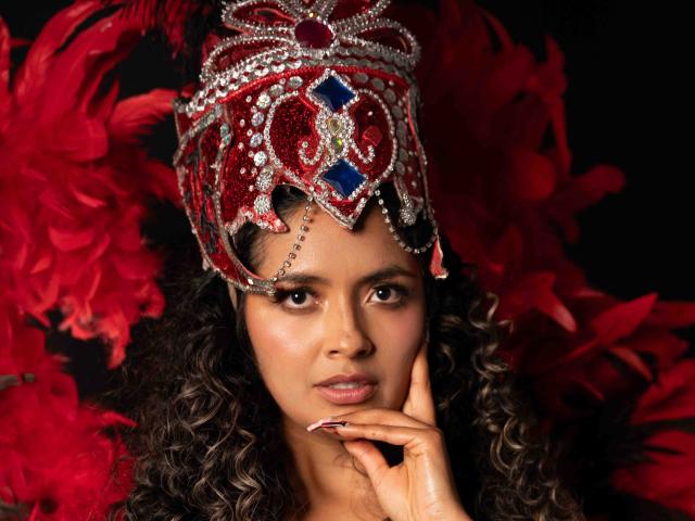 JedaZore lleva una elaborada corona roja y plateada con gemas azules, rodeada de plumas rojas. Ella se toca la barbilla con una mirada seductora, tiene el cabello rizado y oscuro, y una expresión sensual..