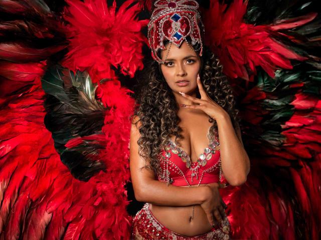 JedaZore, met lang krullend haar, draagt een rood en zilveren bikini en hoofdtooi versierd met pailletten, en heeft grote rode en zwarte veren. vleugels, die haar gezicht raken, een verleidelijke, intense uitstraling, diep rood en zwart..