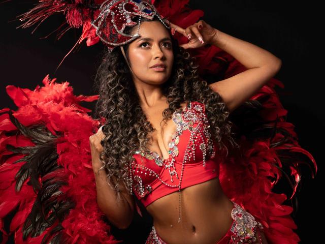 JedaZore, vestida con un top corto rojo brillante y una diadema a juego, estaba rodeada de plumas rojas. Cabello negro, largo y rizado, piel oscura, expresión sensual, mano levantada hacia la cabeza, atractivo sexual intenso..