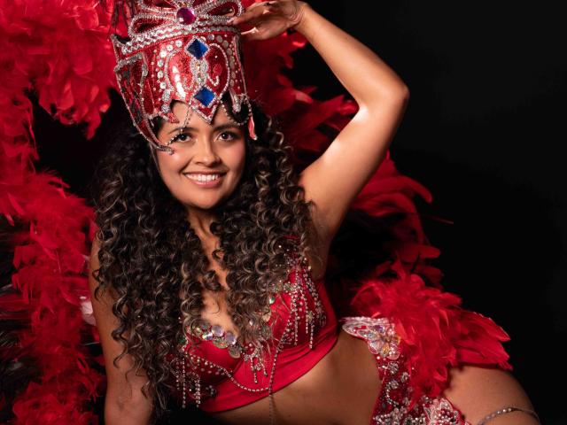 JedaZore, con cabello rizado oscuro, sonriendo, vestida con un traje rojo y plateado adornado con lentejuelas, sosteniendo un tocado a juego. rodeado de plumas rojas. Pose seductora, sexy, confiada, colores vibrantes..