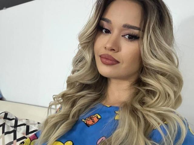 MistyReeds, with long wavy blonde hair, wearing a blue shirt with cartoon characters, gazes directly at the camera with a seductive smirk, full lips slightly parted, heavy makeup. Fucking sexy.