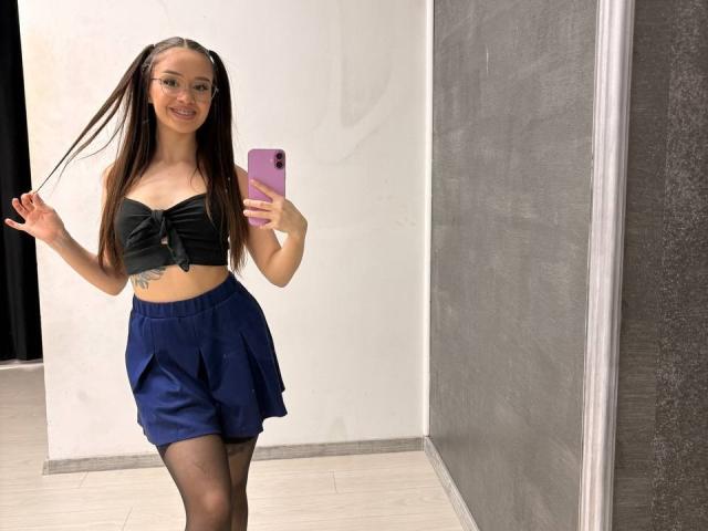 MistyReeds maakt een selfie, staand in een deuropening, gekleed in een zwart topje zonder bandjes en een marineblauwe rok, terwijl ze een roze de telefoon in haar rechterhand. Lang, nat haar, witte achtergrond.