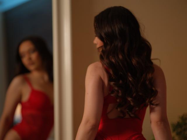MistyReeds stands in a dimly lit room, facing a mirror. She wears a red, lacy lingerie set, showing her back with long, wavy black hair. Her reflection gazes back. Sexual tension is evident.
