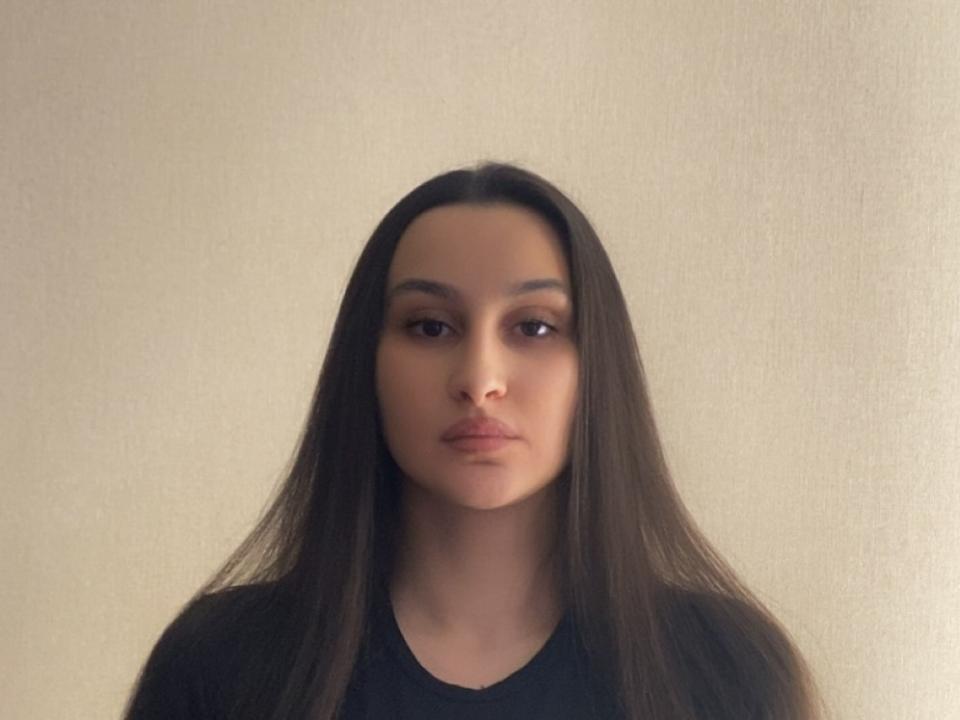 Fotografia di PamDeadmond con lunghi capelli neri lisci, espressione neutra e pelle marrone chiaro, che indossa un abito nero. camicia. Sfondo chiaro. Sguardo concentrato, trucco leggero, leggera ombra sul lato destro..