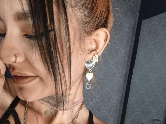 Close-up van LiiaSmith, met bruin haar in een rommelige knot, zwarte pony, neusring, hartvormige oorbellen, zilveren choker. Tatoeage op de nek, bleke huid, lichte grijns, donkere lipstick, grijze achtergrond. Heel aantrekkelijk..