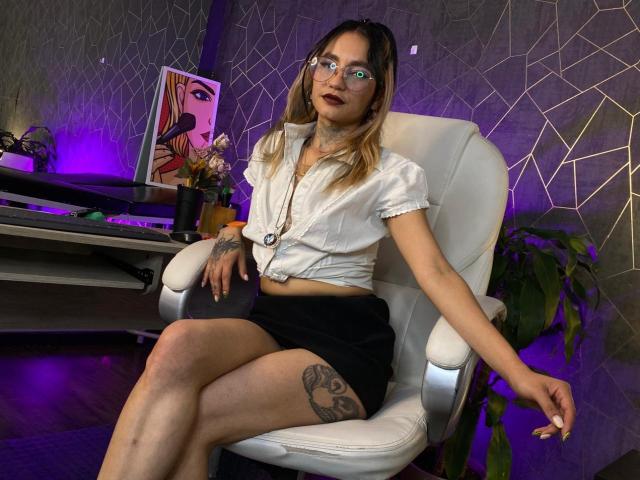 LiiaSmith, usando óculos e uma camisa branca de botões amarrada na cintura, está sentada em uma cadeira branca com as pernas cruzadas. revelando uma tatuagem na coxa. Pênis visível em shorts pretos. Arte de parede neon..
