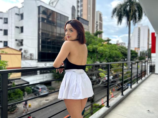 Lara Harvey, de costas, apoiada em uma grade preta, vestindo uma blusa preta sem alças e uma saia branca, mostrando sua silhueta arredondada, nádegas nuas. Cenário urbano com edifícios e palmeira. Incrivelmente atraente..