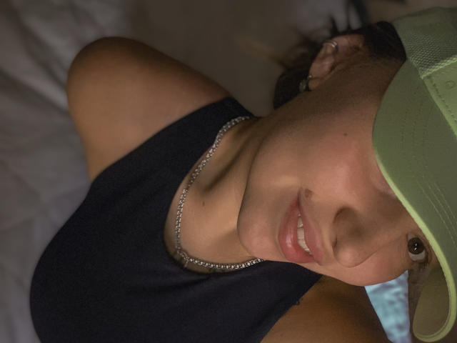 Autorretrato de LoriSue em close-up, com pele morena, usando um boné verde e uma camisa preta, deitada em uma cama, com um colar de prata. sorriso relaxado, iluminação suave, tensão sexual sutil, ambiente descontraído..