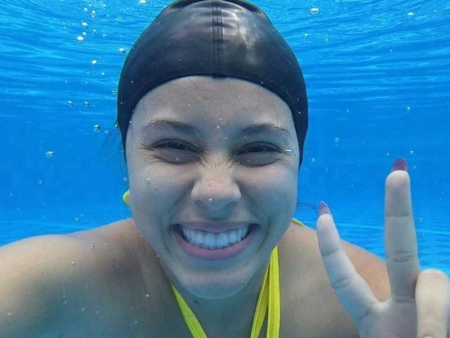 Underwater photograph of MarilynHills with a wide, smiling face, eyes squinted shut, wearing a black swim cap and yellow bikini top. She gives a peace sign with her right hand. Blue water background.