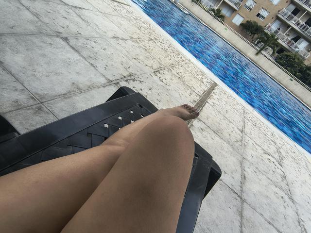 Close-up of MarilynHills' legs crossed at the knees, feet resting on a black bag near a blue-tiled pool. Light skin tone, smooth texture. Background includes blurred poolside structures.