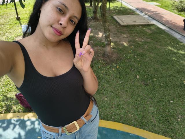 Photograph of MarilynHills, a Latina woman with long black hair, wearing a black tank top and blue jeans with a brown belt, flashing a peace sign, outdoors on green grass.