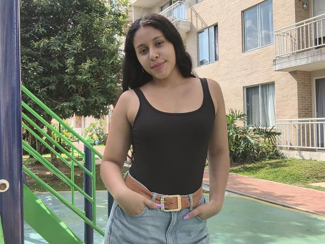 MarilynHills, medium build, tan skin, black tank top, light blue jeans, brown belt, hands in pockets, standing by green metal railing, apartment building background, hot summer vibe, casual look.