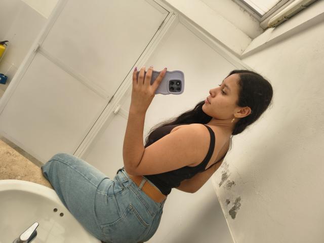 MarilynHills kneels in a bathroom, taking a mirror selfie with a smartphone. She has long black hair, a black tank top, and tight blue jeans. She's looking at the phone with a sultry expression.