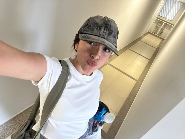 SpencerLe prend un autoportrait dans un couloir bien éclairé. Elle porte une casquette plate grise et un t-shirt blanc, avec un sac bandoulière vert et des objets bleus en arrière-plan. Pose décontractée, de la taille d'un coq..