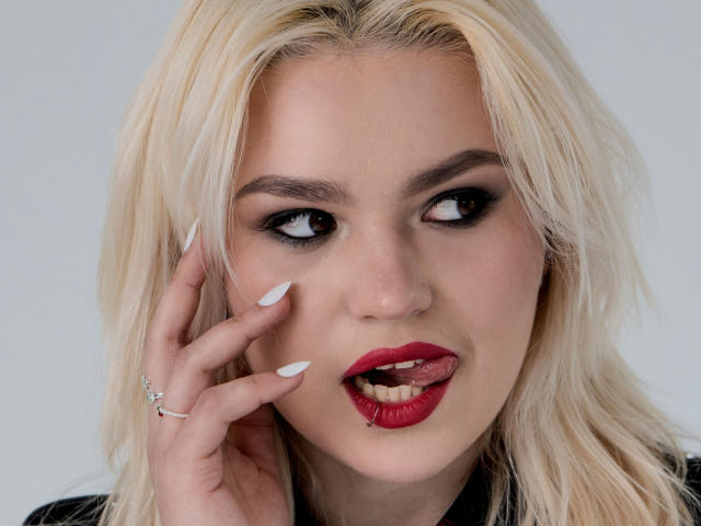 Primer plano de SweetLilyy, una mujer rubia con lápiz labial rojo, uñas blancas y un anillo de plata en su mano derecha. Está mordiéndose el labio y mirando hacia un lado con una expresión seductora..