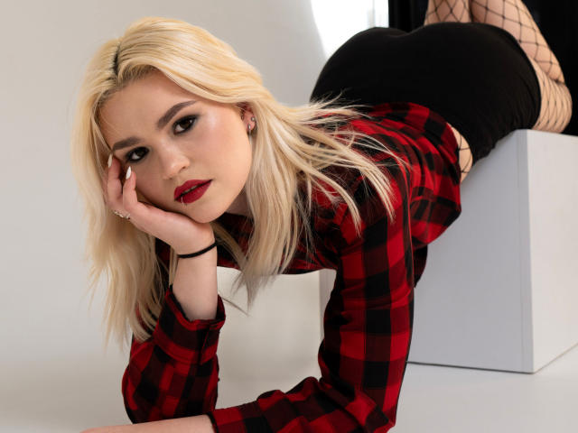 Fotografia di SweetLilyy con capelli biondi, camicia a quadri rossa, minigonna nera, collant a rete, in posizione leggermente inclinata in avanti. blocco bianco, mano sul viso, rossetto rosso, sguardo intenso, tensione sessuale, posa seducente.