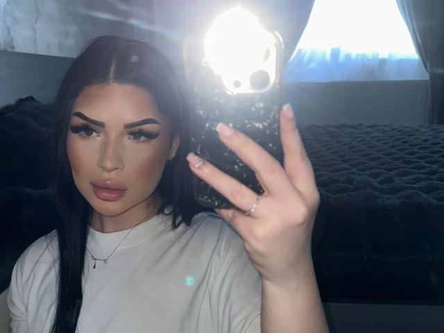 Foto van Meryshor met lang zwart haar, veel make-up en volle lippen, die een telefoon vasthoudt met een fel licht. Een wit T-shirt aan, hand omhoog, sexy uitdrukking, directe blik..