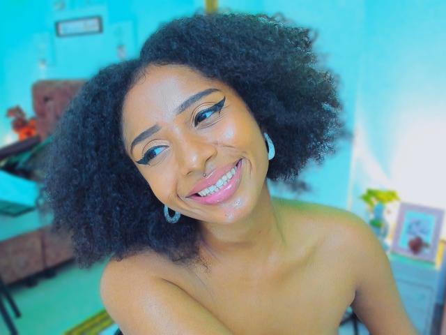 Primer plano de Megan Williams, con piel de color marrón medio, cabello negro rizado y una amplia sonrisa. Está sin sujetador, mostrando sus hombros. Fondo azul brillante, muebles borrosos y una planta amarilla. Muy atractivo..