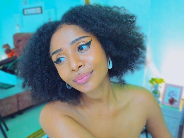 Close-up de Megan Williams, uma mulher negra com cabelo cacheado natural, usando delineador de asa marcante e batom rosa. Deitada de bruços, olhando diretamente para a câmera com um leve sorriso. Fundo azul claro.