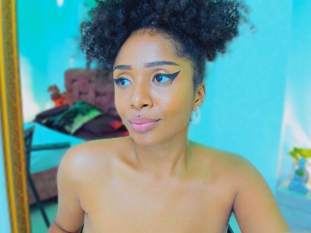 Close-up of MeganWilliams with dark skin, curly black hair, and blue eyeshadow. She's topless, with a slightly seductive gaze, and a subtle smile. Bright blue background, blurred furniture.
