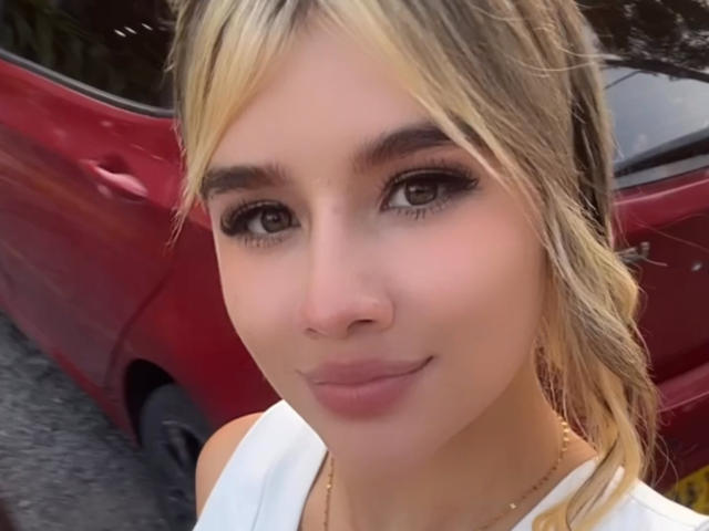 Close-up photo of SashaPolet, blonde hair in a ponytail, wearing a white top, standing in front of a red car. She has large, dark eyes, heavy eyeliner, and a slight smile. Fucking hot.