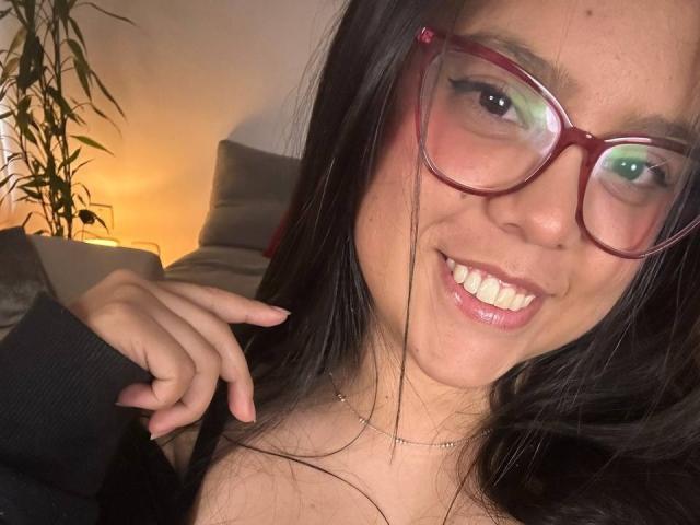 Close-up selfie of ConnySaenz, a smiling woman with long black hair, red-framed glasses, and visible teeth. She's wearing a black top, looking directly at the camera with a flirty expression.