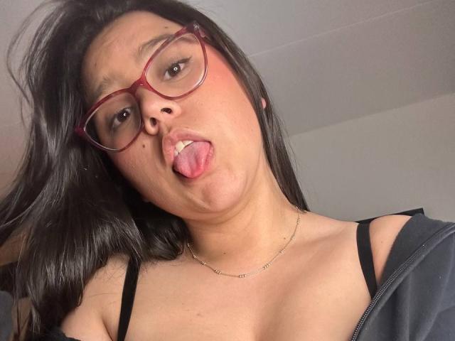 ConnySaenz has long black hair, red glasses, and a black top with straps. She's sticking out her tongue with a teasing expression, looking directly at the camera. Her neck has a thin chain.