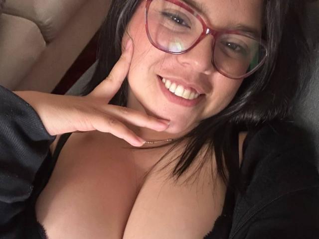 Close-up of ConnySaenz, a woman with large breasts, wearing red glasses and a black top, smiling seductively, hand touching her neck, visible cleavage, casual photo with sexual tension.