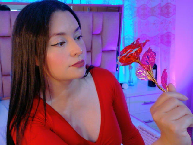 VaaleryStark mit langen, schwarzen Haaren, die ein rotes Top mit V-Ausschnitt trägt und rote und rosa Blumenstiele hält, blickt zur Seite. Ein sinnlicher Ausdruck, in einem hell erleuchteten Raum..