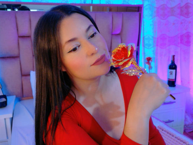 VaaleryStark, con cabello largo y oscuro, vestida con una blusa roja, sosteniendo una flor roja y amarilla, lamiéndola de forma seductora. Luces azules y moradas brillantes, una botella sobre una mesita de noche..