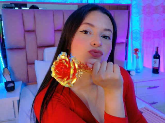 VaaleryStark, una giovane donna con lunghi capelli neri, tiene vicino al viso una rosa rossa luminosa, indossando una maglietta rossa, in un ambiente molto luminoso. Camera da letto luminosa. Le luci blu e viola esaltano l'atmosfera..