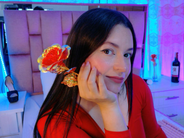 VaaleryStark, con i suoi lunghi capelli neri, indossa una camicia rossa e una clip a forma di pesce colorata, appoggiando il mento sulla mano, su uno sfondo blu. illuminazione al neon.
una bottiglia è appoggiata su una scrivania dietro di lei.