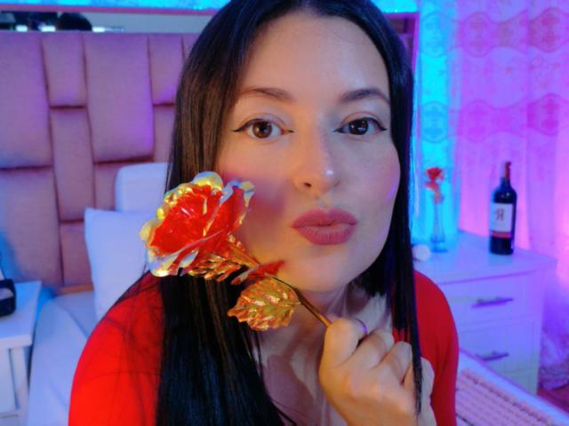 VaaleryStark, una mujer asiática con cabello negro largo, labios rojos, sosteniendo una flor roja y amarilla cerca de su rostro, vestida de rojo. camisa, dormitorio con iluminación azul y púrpura, botella de vino en el fondo.