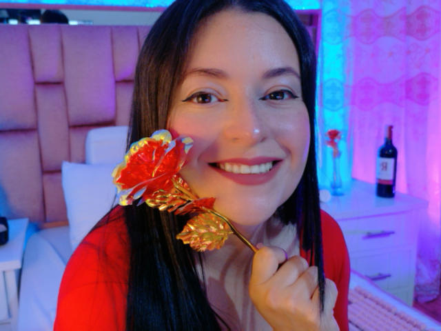 VaaleryStark, con cabello largo y negro, sonriendo, sosteniendo un caramelo de color rojo y naranja cerca de su boca. Ella lleva una camisa roja y está sentada en un dormitorio muy iluminado. Increíblemente atractivo/a..