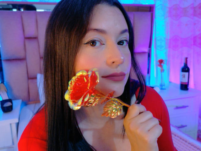 VaaleryStark, with long black hair, holds a red and gold foil-wrapped lollipop to her lips. She wears a red top. Blue and purple lighting. Bottle on a table in background.