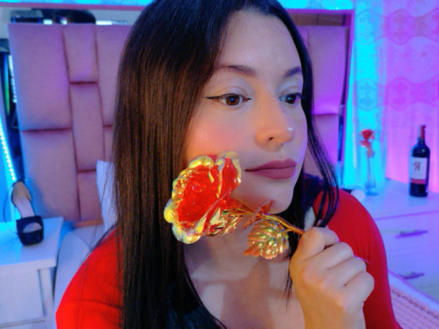 VaaleryStark, with long dark hair, holds a glowing red and gold flower to her lips, wearing a red shirt, in a neon-lit bedroom, bottle on a table in the background.