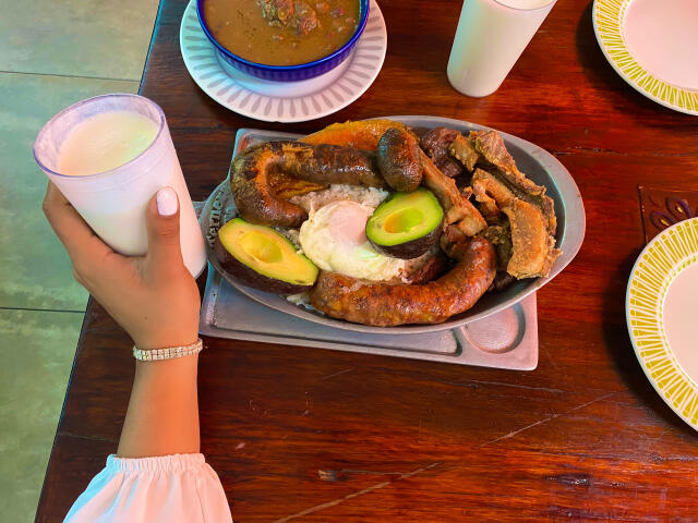 Un primer plano de una mano sosteniendo una taza blanca sobre un plato de salchichas a la parrilla con rebanadas de aguacate y una cucharada de crema. Mesa de madera oscura, dos platos de papel.