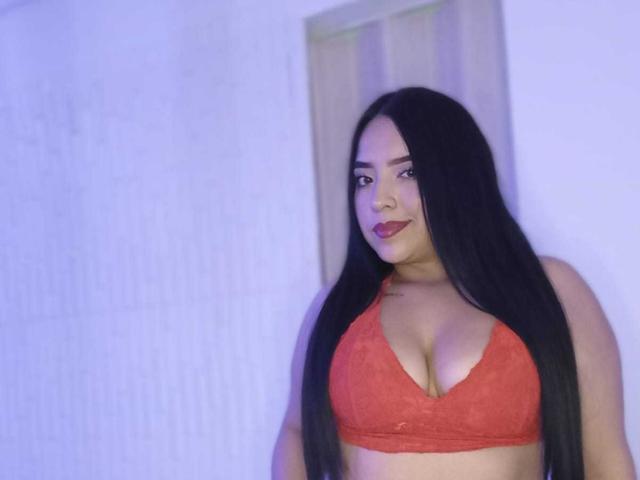 Photograph of AdaraCute69, a curvy woman with long black hair, wearing a red lace bra that accentuates her large breasts. She has a confident expression, with blue lighting in the background.