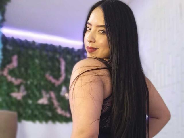 AdaraCute69, with long black hair, turns back to look over her shoulder, wearing a black lace top, dark red lipstick, and a slightly seductive smile. Green wall in the background.