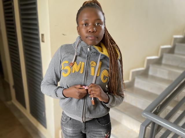 Yollah, uma mulher negra com longas tranças, vestindo um moletom cinza com zíper e as letras "SMC" em amarelo, está em pé nos degraus de uma escada. segurando um objeto rosa, olhando diretamente para a câmera, expressão séria, pose casual.